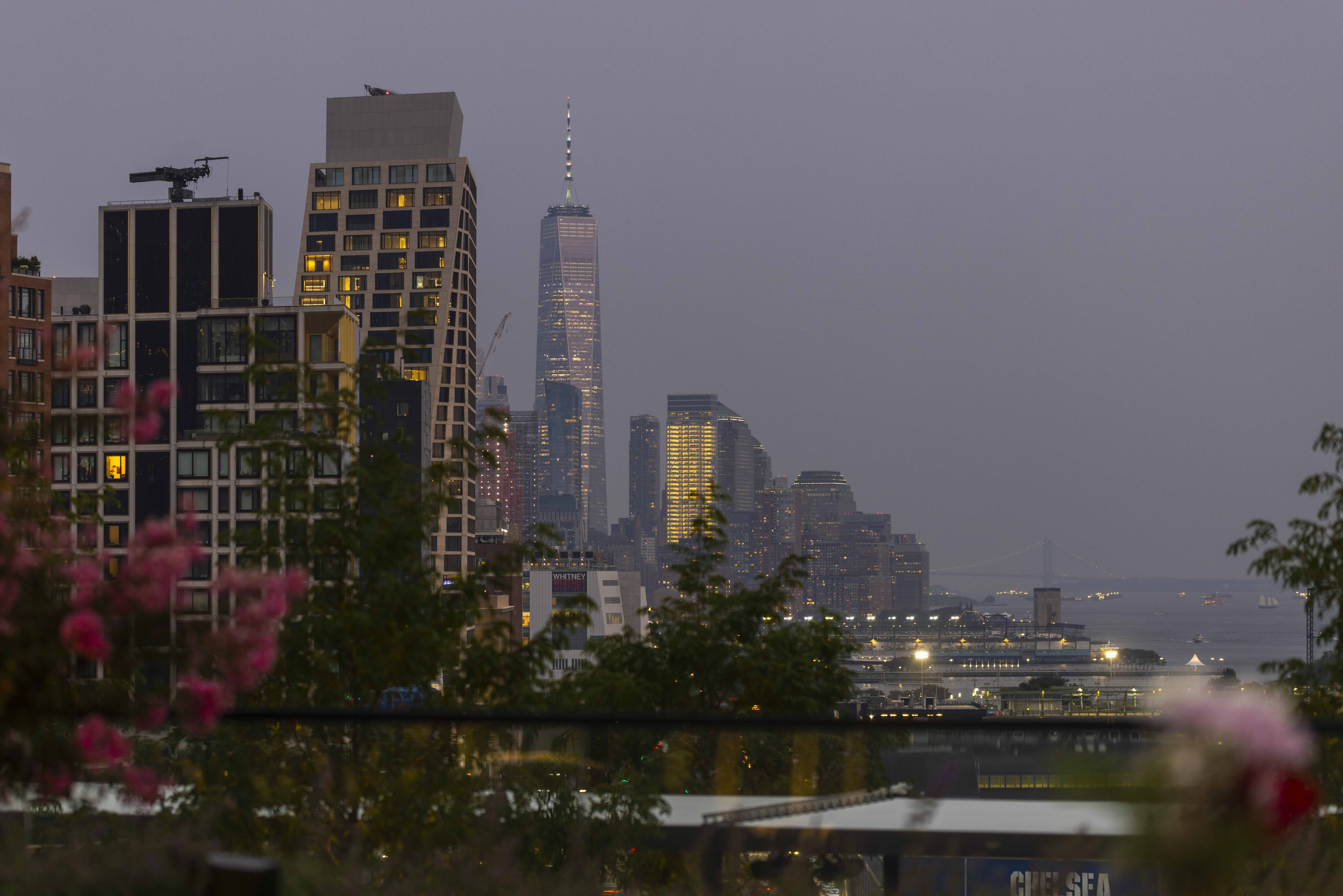 Evening cityscape with one world trade center in the background, framed by buildings, flowers & a ferry on the hudson river.