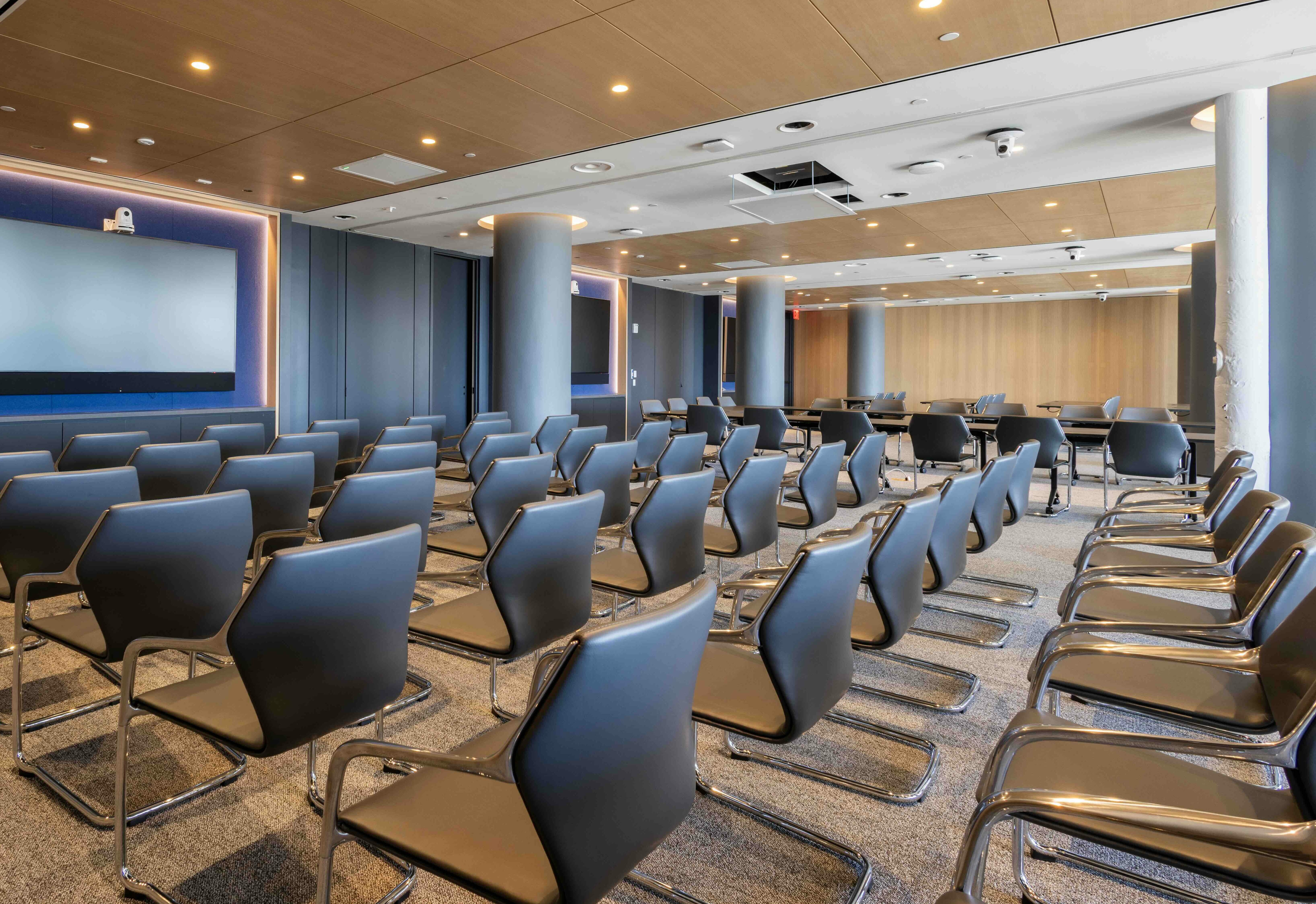 Modern seminar room with rows of sleek black chairs facing a large screen, featuring wood-paneled ceiling & ambient lighting.