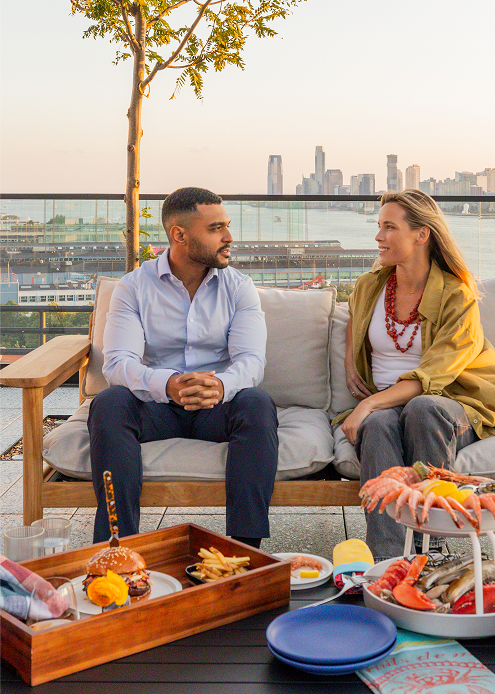 Two guests dining on level10 rooftop, sharing seafood and burgers with city and water views behind them.
