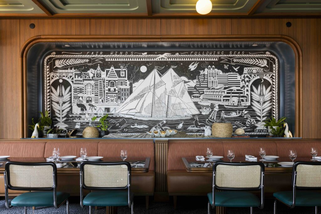 Stylish restaurant interior with nautical mural, featuring a sailing ship and coastal scenes above dining tables and decor.