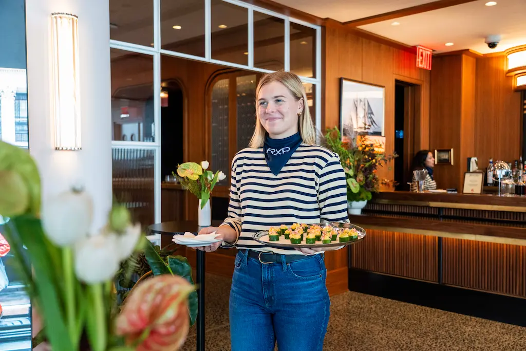 Smiling server in striped shirt holding tray of hors d'oeuvres in elegant lobby with wood paneling, flowers & nautical decor.