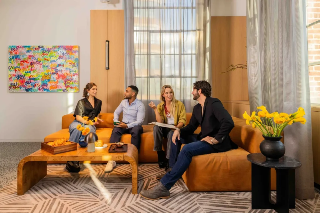 Four people chatting in a cozy living room with leather sofas, a wooden coffee table, and a colorful inspirational art piece.