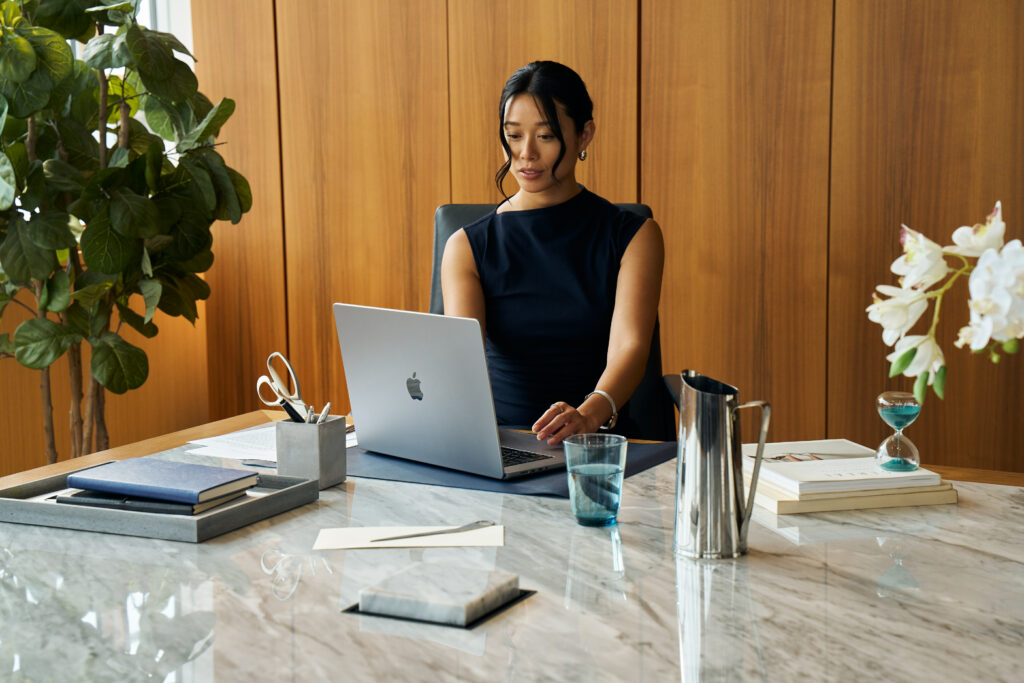 Woman working on a laptop at a marble desk in a modern office with wood paneling, stylish decor, and natural light.