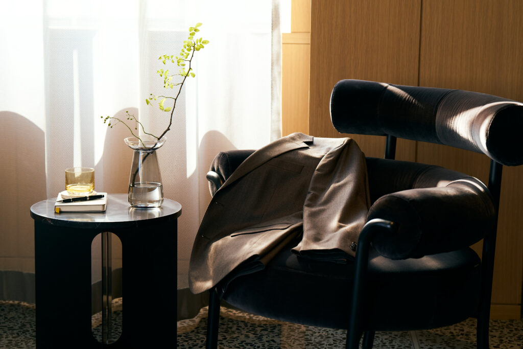 Cozy lounge corner with black velvet chair, beige blazer draped over arm, and side table with drinks, books & a leafy branch.