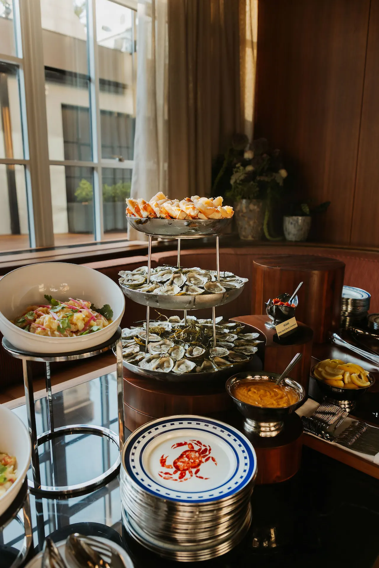 Seafood buffet display featuring oysters, crab legs, sauces, and salads arranged elegantly near a sunlit window.