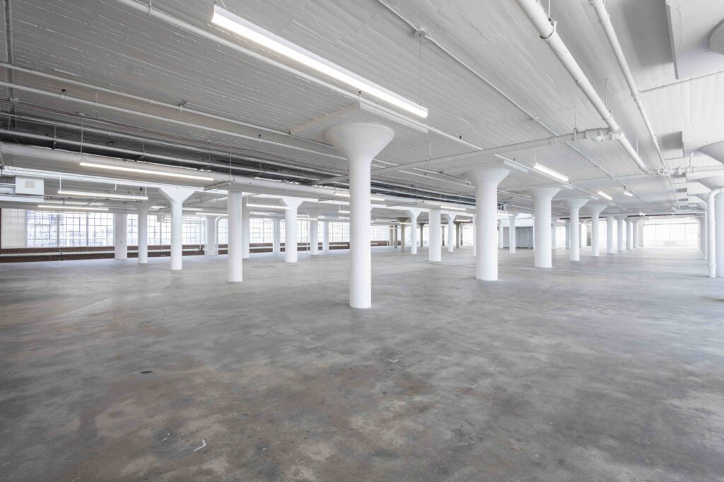 Empty industrial space with concrete floors, white columns, exposed piping, and large windows letting in natural light.