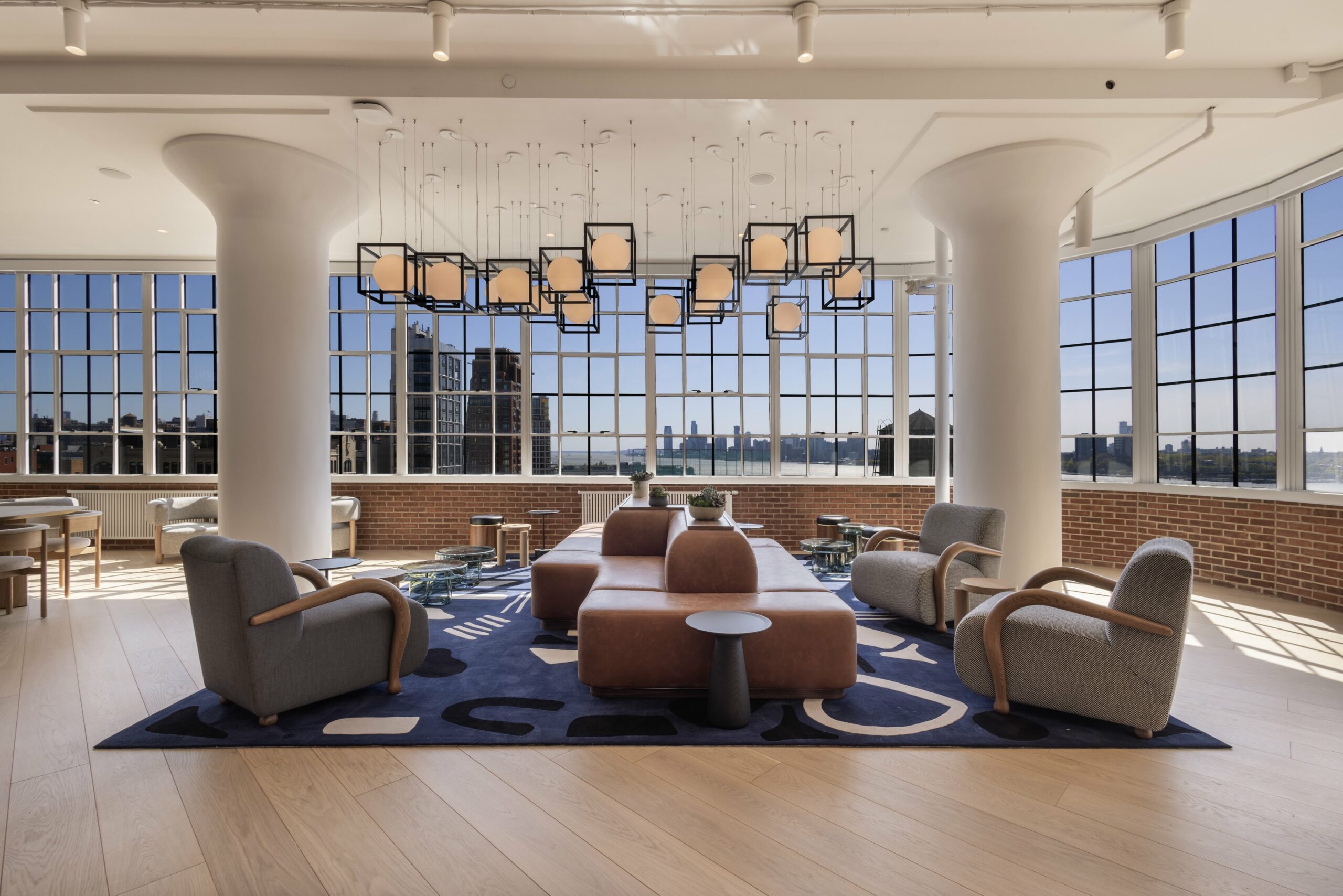 Lounge with panoramic city views, featuring armchairs, a leather sectional sofa, abstract rug, and geometric chandelier.