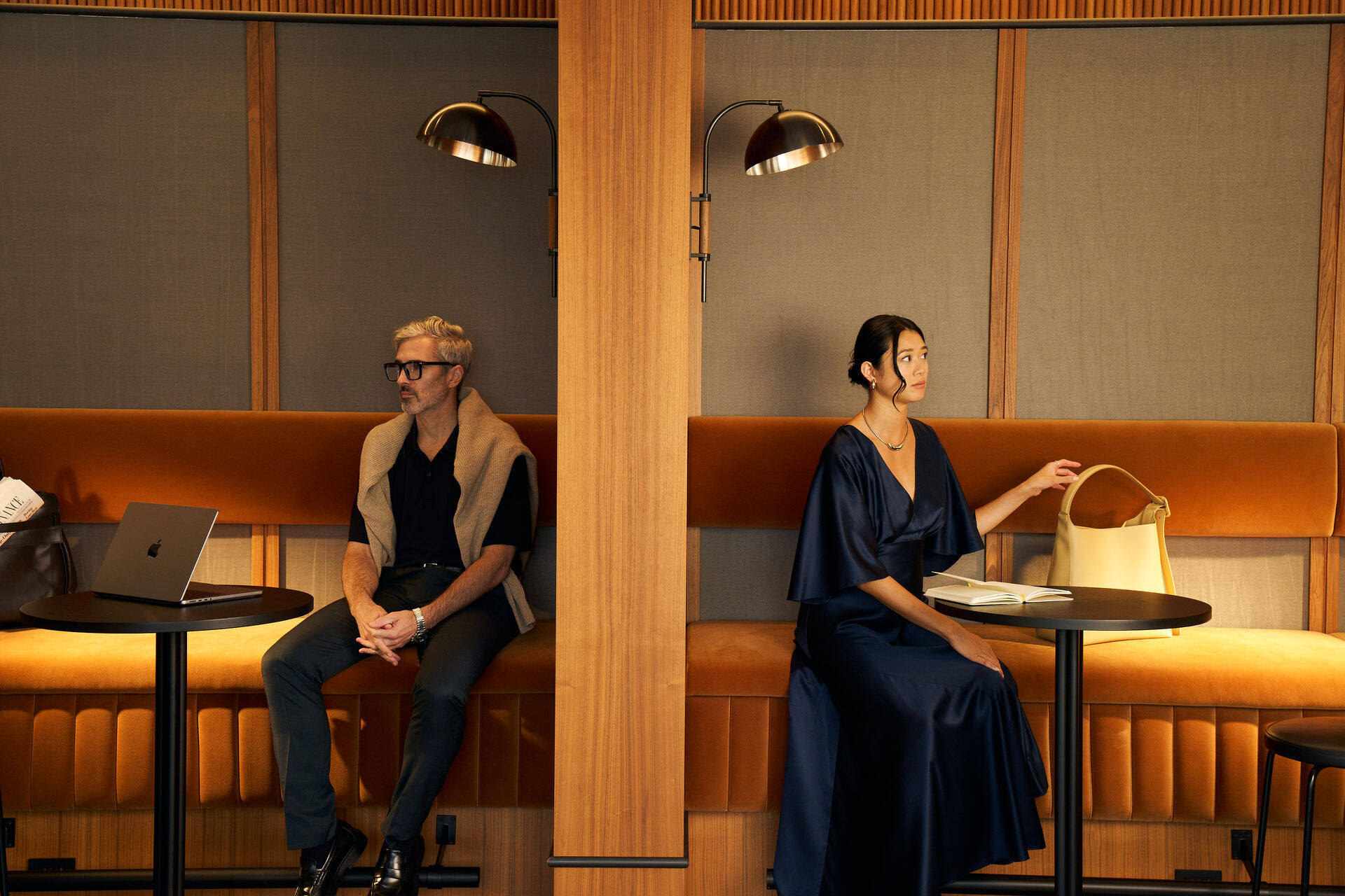 Two professionals seated in adjacent private lounge booths within a sophisticated hospitality workspace, featuring warm wood paneling, amber velvet banquettes, modern task lighting, and intimate seating areas designed for remote work, meetings, and quiet relaxation.