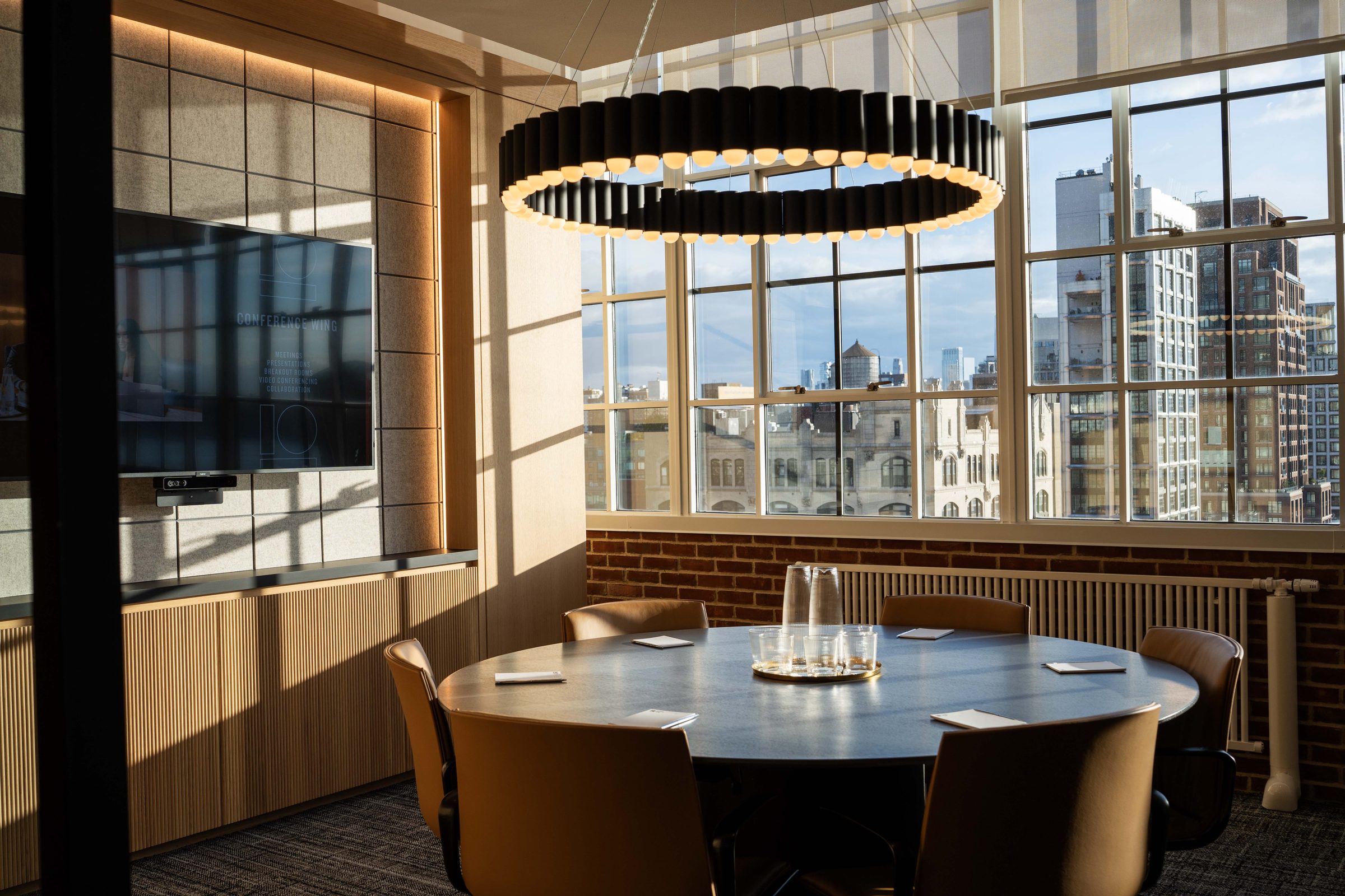 Intimate conference room at level10's conference wing with a round dark wood table, caramel leather chairs, a large display screen on an acoustic wall panel, circular chandelier pendant light, and city skyline views through gridded windows