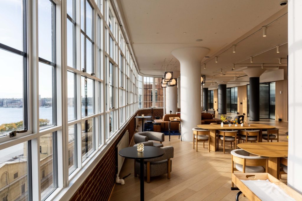 Upscale lounge at level10 with floor-to-ceiling windows overlooking the hudson river and new jersey skyline, featuring cognac leather sofas, globe pendant lights in black geometric frames, and exposed brick walls