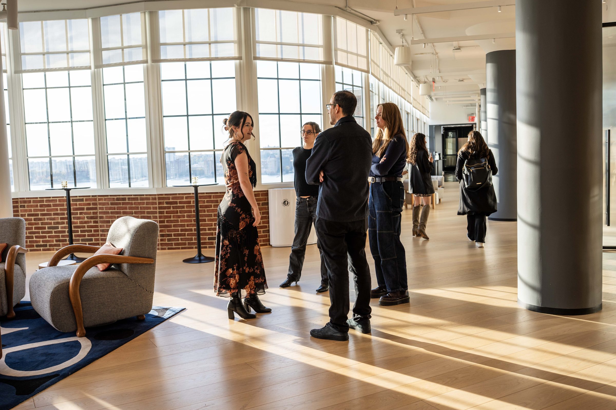 Panoramic view of Level10 riverside lounge seating area with cognac leather sectional sofa, globe pendant chandeliers, exposed brick wall, and sweeping Hudson River views at sunset through industrial-style windows